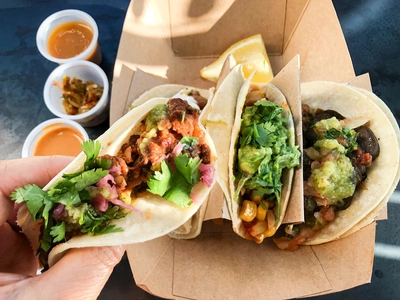 Drei Tacos in einer Pappschale, gefüllt mit Fleisch, Bohnen, Avocado, Salsa und frischem Koriander. Eine Hand hält einen Taco, während im Hintergrund drei kleine Schälchen mit Saucen und Zitronenscheiben auf einem dunklen Tisch stehen.