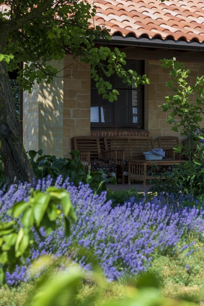 Ein blühendes Lavendelfeld erstreckt sich vor einem Haus mit rotem Ziegeldach und einer schattigen Veranda. Unter einem großen Baum steht eine hölzerne Sitzgruppe mit Stühlen und einem Tisch. Sonnenlicht fällt durch das Laub und beleuchtet die violetten Blüten.